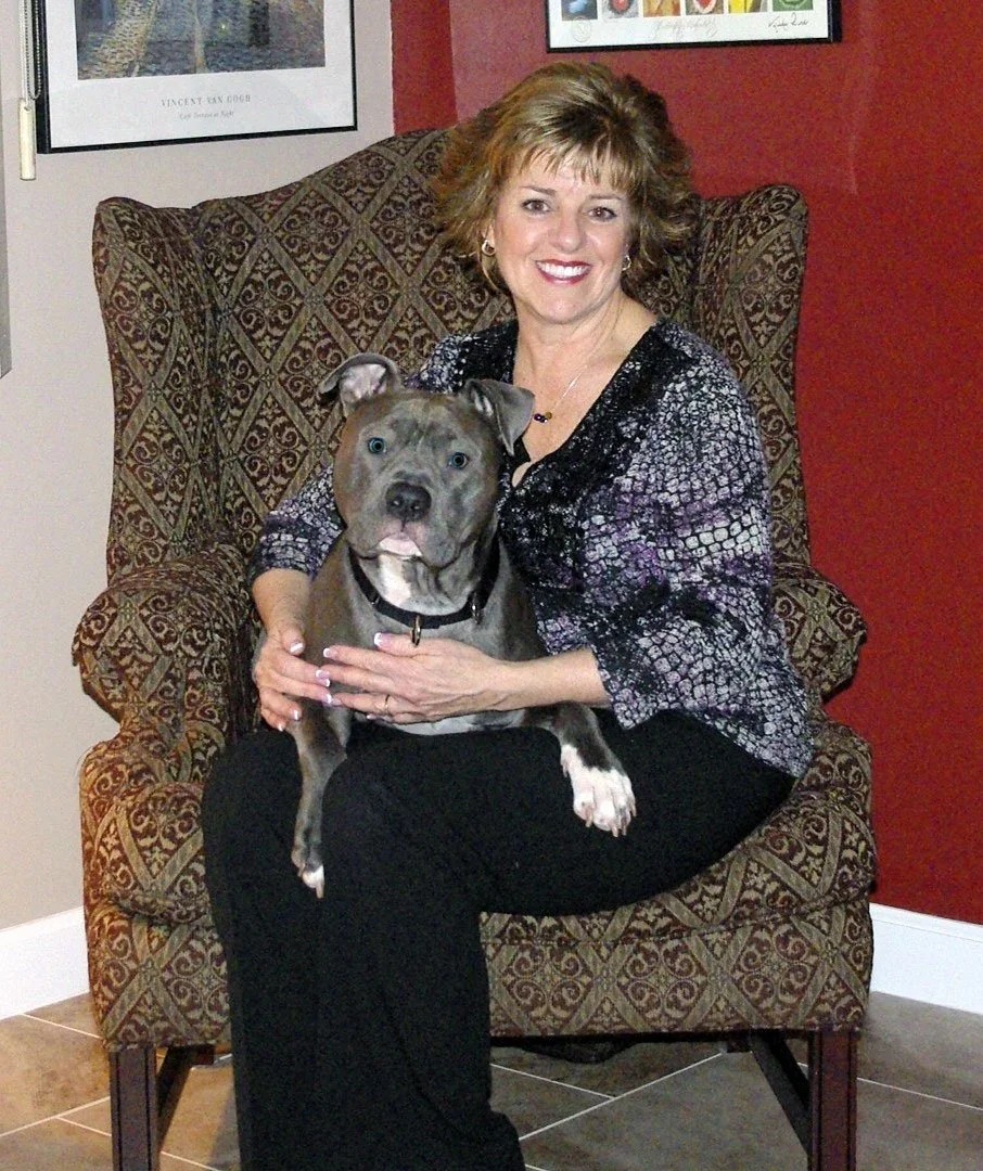 A woman sitting in a patterned armchair holding a gray pitbull dog on her lap, both smiling at the camera in a room with red and white walls.