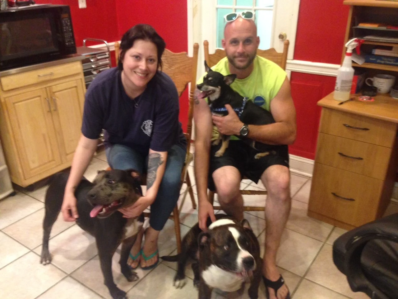 A woman and a man with three dogs in a kitchen. The woman is squatting, petting a black dog with a tan face, and the man is sitting, holding a small black and tan dog. There is a large black and white dog sitting on the floor between them. The kitche