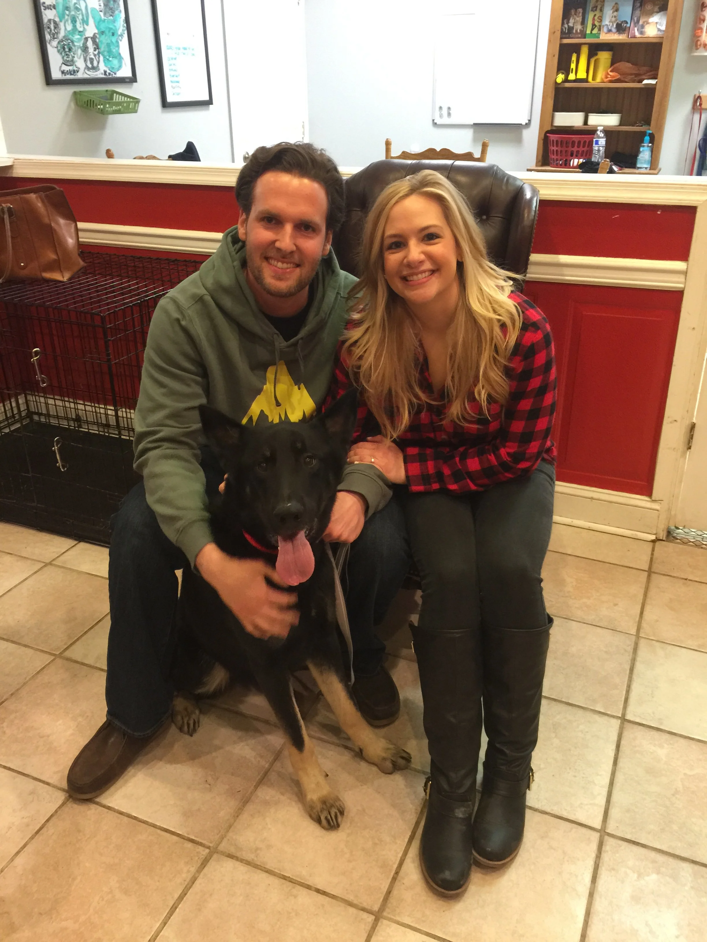 A smiling man and woman sitting on the floor with a large black German Shepherd dog. The man has dark hair and a beard, wearing a green hoodie. The woman has long blonde hair, wearing a red and black plaid shirt and black boots. They are in a room wi