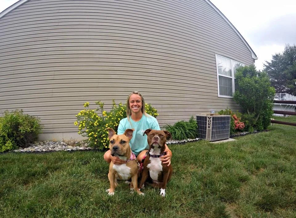 A woman kneeling on the grass, smiling, with two dogs in front of her. One dog is tan and white, and the other is brown with white markings. They are outdoors in a backyard with a gray house, bushes, and an air conditioning unit in the background.