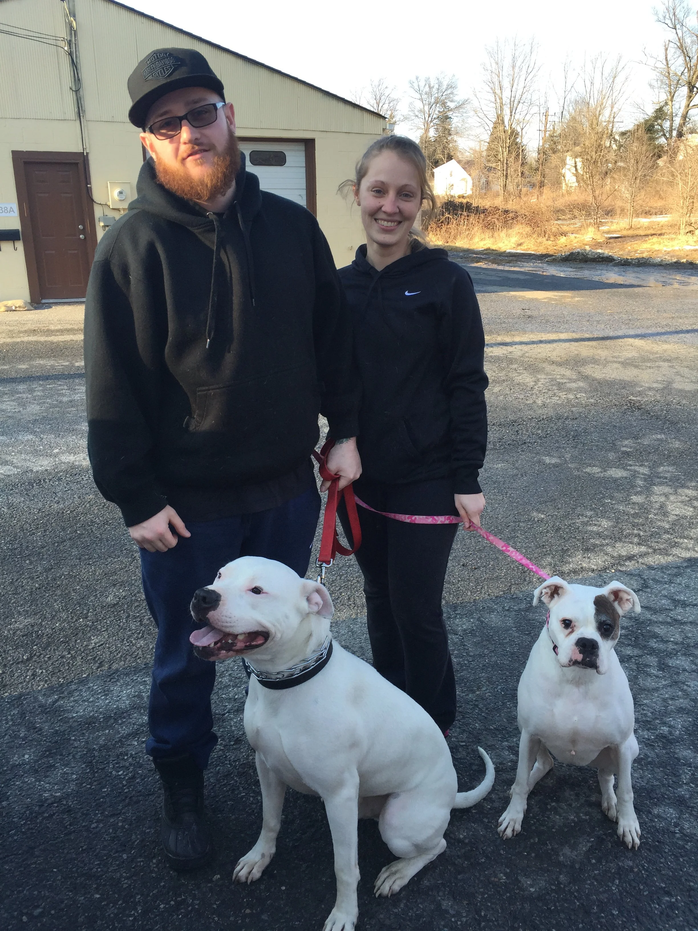 Two people posing outdoors with two large white dogs on leashes. One person is a man with red beard, wearing glasses, a black hoodie, and a black cap. The other person is a woman with light brown hair, wearing a black Nike jacket. The background feat