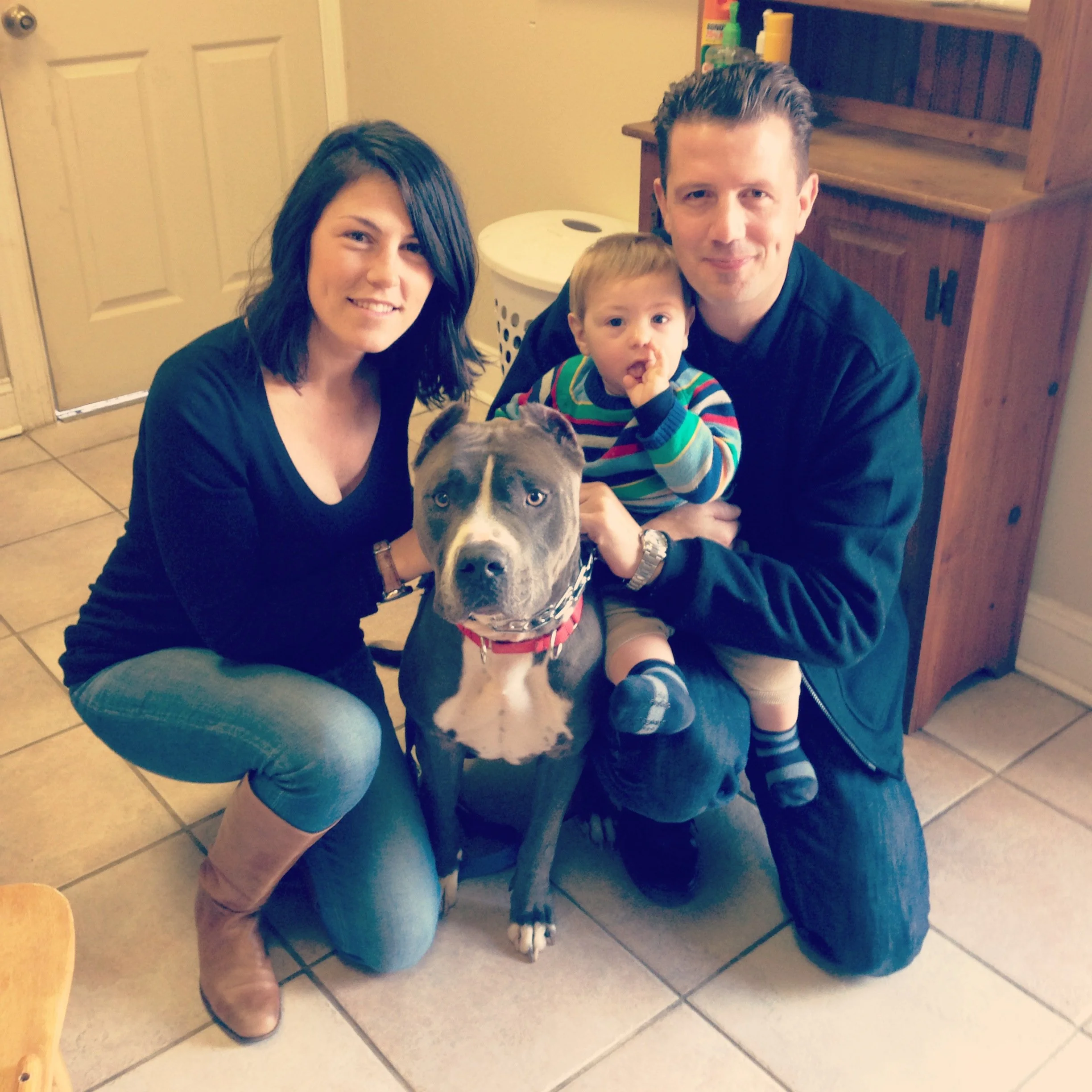 A family of three sitting on the floor in a room, with a woman, a man, a young boy, and a dog in the picture. The woman has shoulder-length dark brown hair, the man has short brown hair, and the boy is wearing a colorful striped shirt. The dog is a l