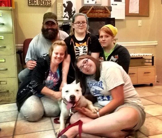 Five people and a white dog sitting and kneeling on the floor smiling indoors, with furniture and posters on the wall behind them.