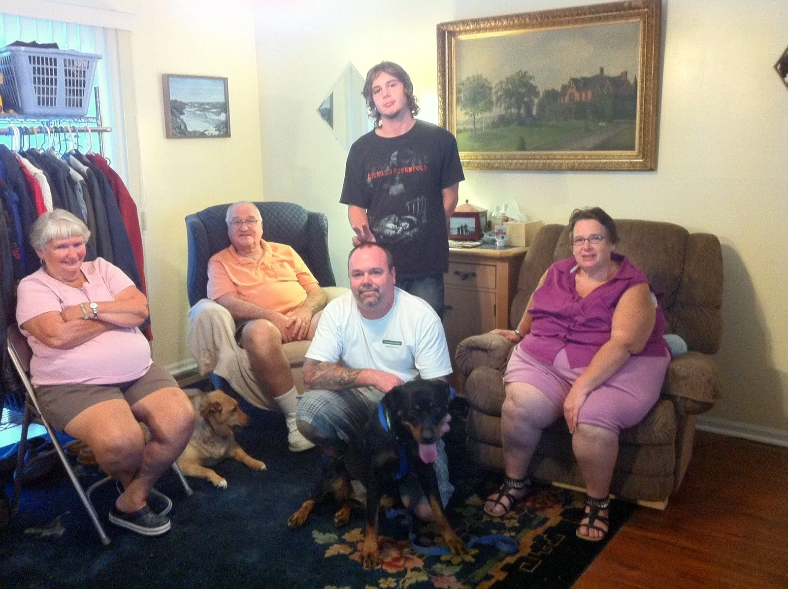 A group of five people and two dogs in a living room. The group includes three elderly women, half of whom are sitting on chairs and one on a sofa, a bearded man with tattoos sitting on the floor, and a teenage boy standing behind them. The room has 