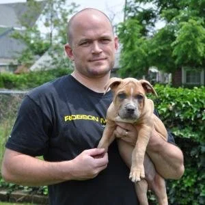 A man with a shaved head wearing a black T-shirt holding a tan puppy in his arms outdoors with trees and houses in the background.