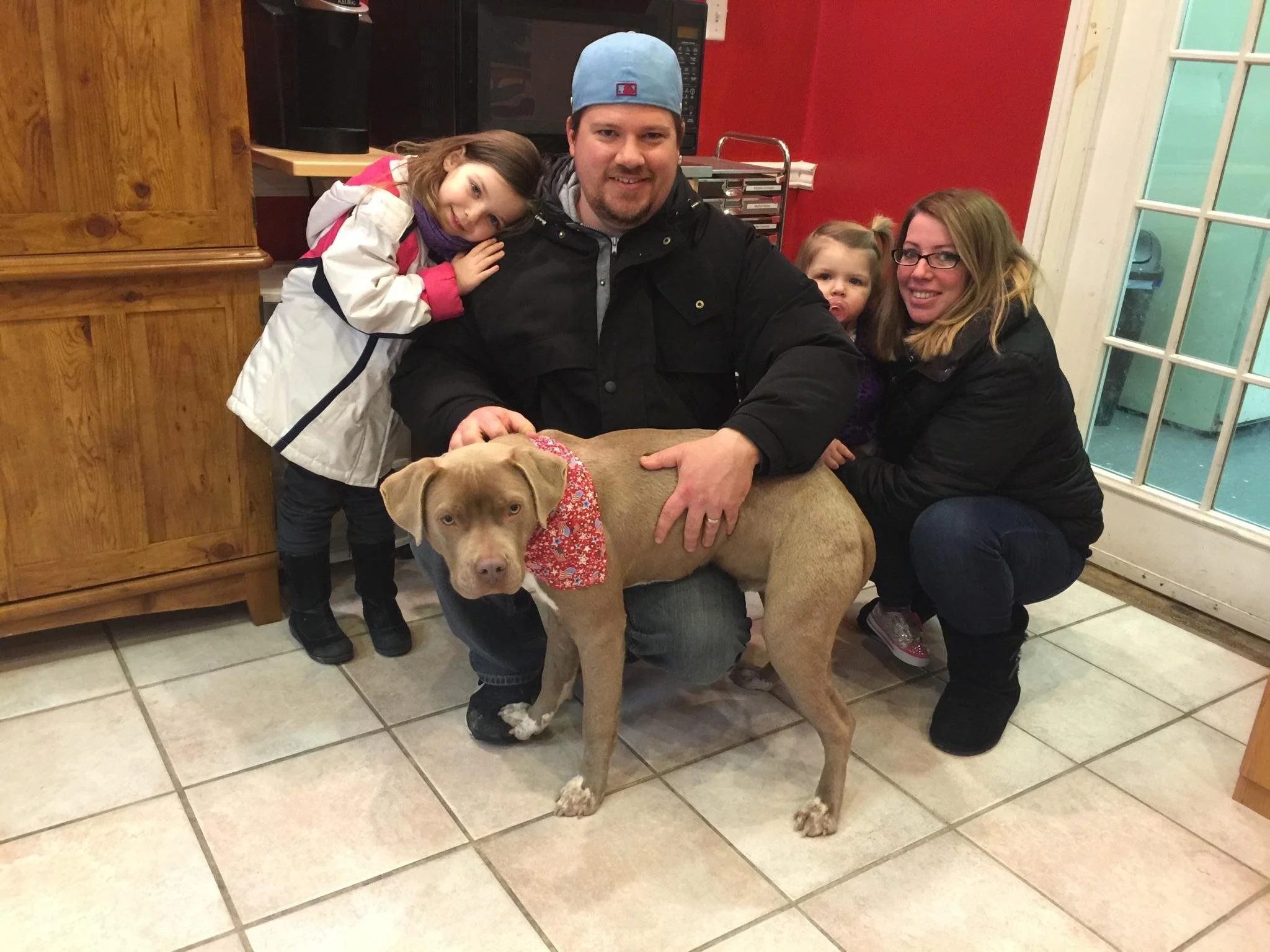 A man wearing a blue cap and black jacket sits at a table with his two young daughters and a large tan dog with a red bandana. The girl on the left leans her head on the man's shoulder, and the girl on the right sits behind the dog. They are inside a