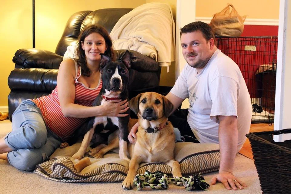 A couple sitting on the floor with two dogs, a woman wearing a red and white striped shirt and ripped jeans, and a man in a white shirt. They are indoors in a living room with a black leather recliner, a dog crate, and various household items visible