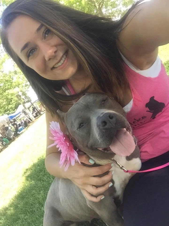 A young woman with shoulder-length brown hair taking a selfie with her arm around a happy, gray pitbull dog wearing a pink flower collar. They are outdoors on a sunny day in a park.