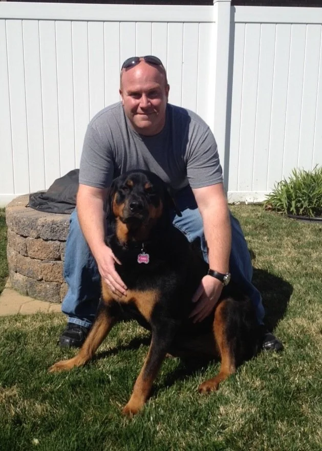 A man crouching in a backyard with a Rottweiler dog.