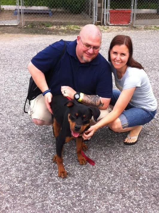 A man and a woman kneeling outdoors on a gravel surface, smiling, with a black and tan puppy between them. The man has a shaved head, glasses, and tattoos, wearing a navy shirt and khaki shorts. The woman has long brown hair, wearing a light gray t-s