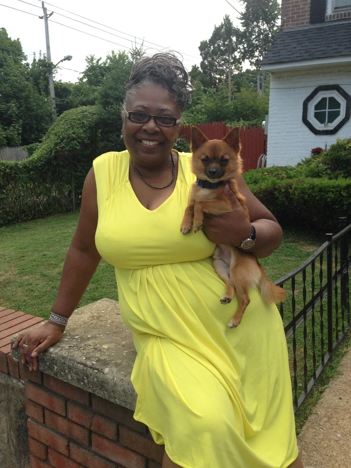 A woman in a bright yellow dress holding a small brown dog in a residential yard with green trees, a white house with black trim, and a red fence in the background.