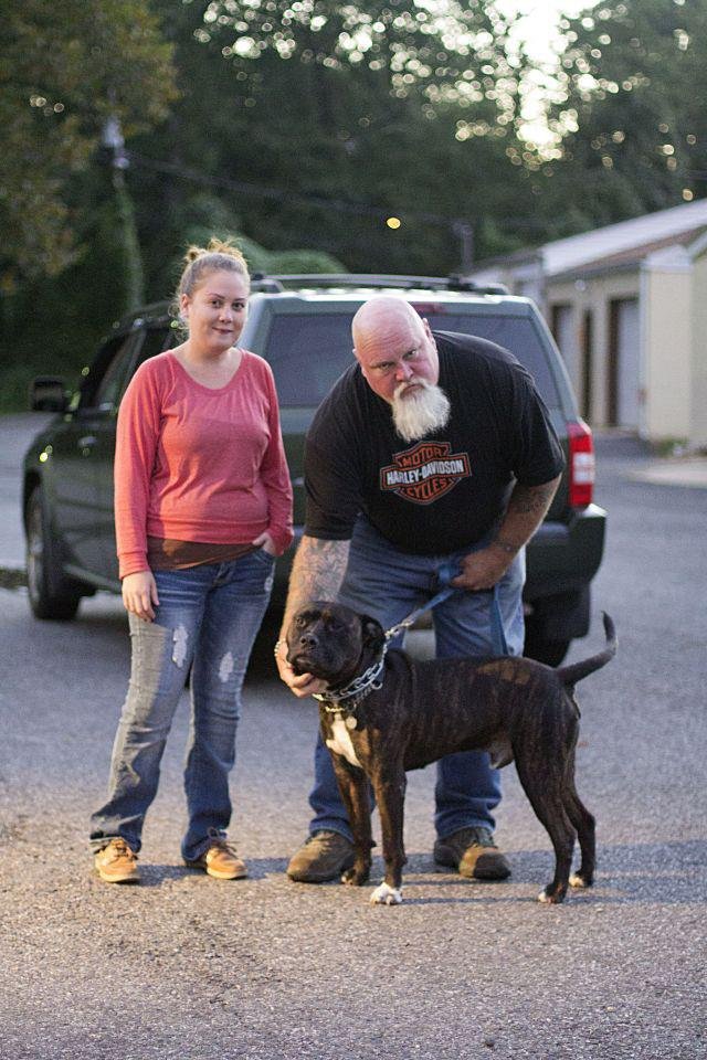 A young woman with a top knot hairstyle and a man with a white beard and tattoos pose with a black dog on a leash on a street with a parked vehicle in the background.