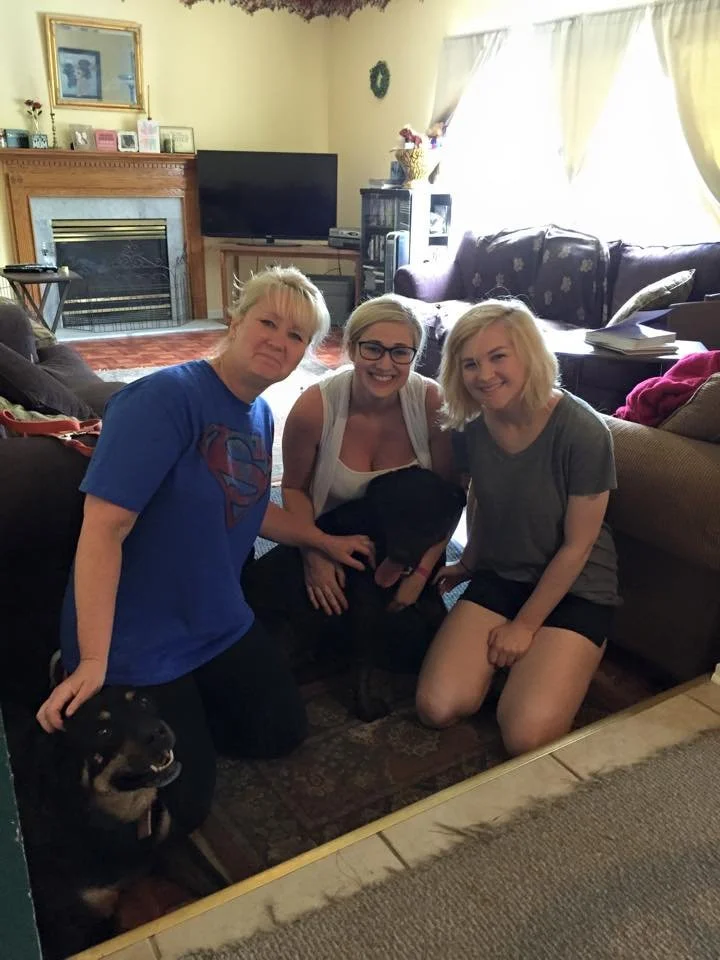 Three women and two dogs in a living room, with a fireplace, TV, and beige curtains in the background.