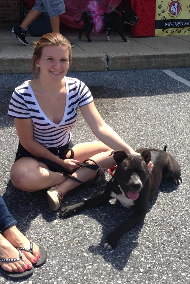 Young woman sitting on the pavement with a black and white dog. The woman is smiling and wearing a striped shirt. The dog is lying down with its tongue out. In the background, there is a small dog on a leash and some colorful banners or signs.