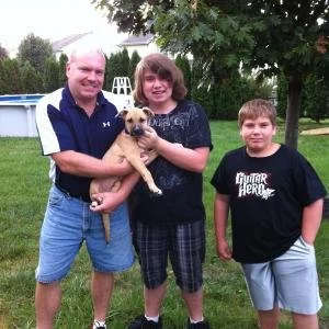 Three smiling people and a puppy outdoors in a backyard, with trees and a house in the background.