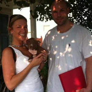 A smiling woman holding a small puppy, standing next to a man outdoors during daytime.