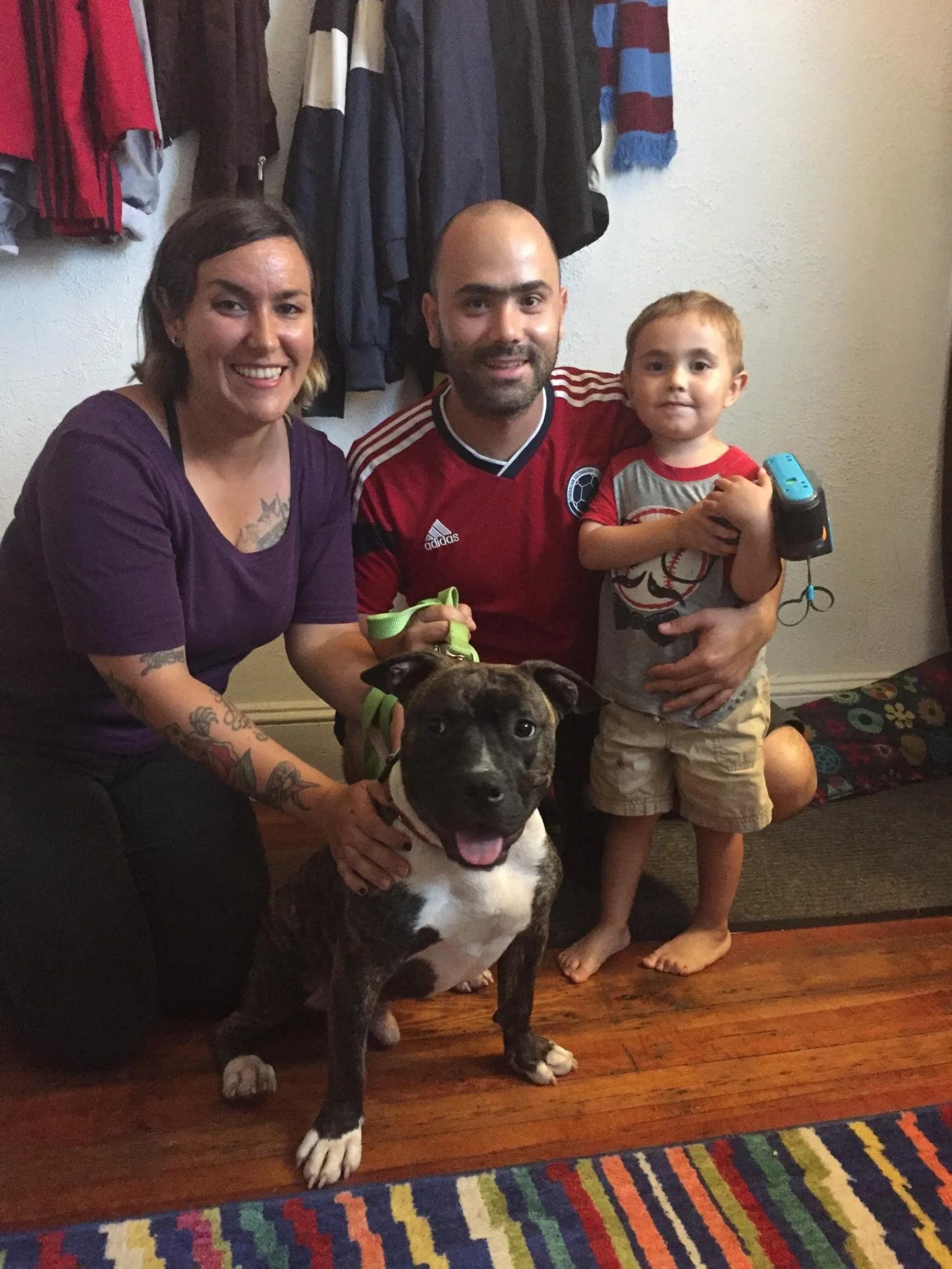 A family photo with two adults, a young boy, and a dog in a room with a colorful rug and hanging clothes in the background.