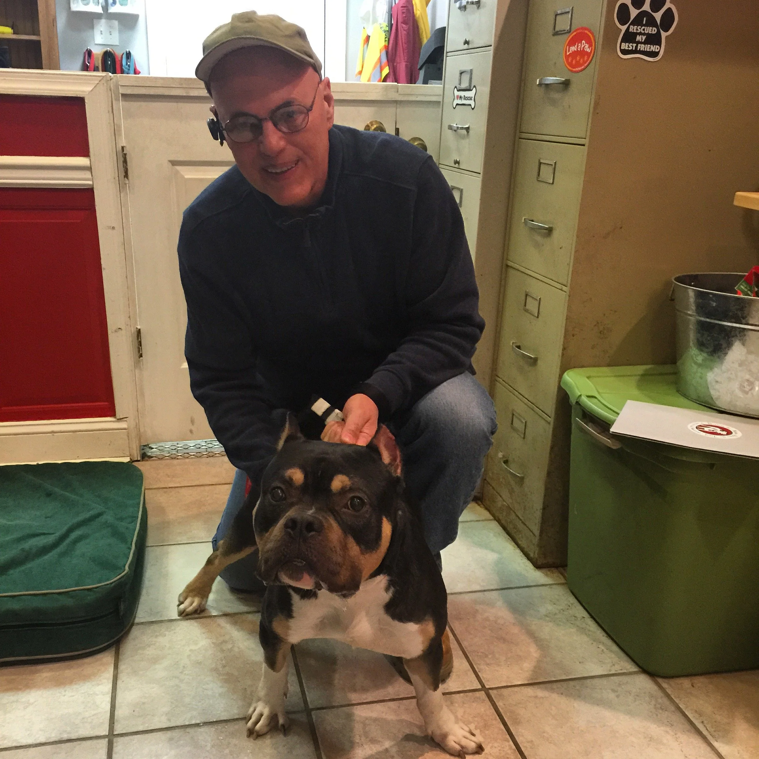 A man with glasses, wearing a tan cap and dark clothing, is smiling and leaning over a large black and tan dog in an indoor setting. The dog is looking directly at the camera with one paw resting on the man's lap. The background features lockers with