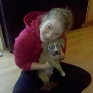 A girl with curly hair wearing a red hoodie, smiling and hugging a small brown puppy with a dark face inside a room with wooden flooring.