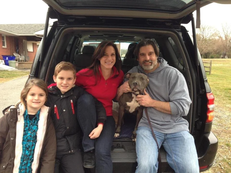 Family of four with a dog, sitting on the open trunk of a black SUV outdoors during daytime.