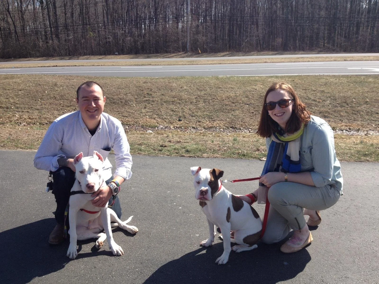 Two people, a man and a woman, kneeling on a paved road holding two dogs. The man has short hair, is wearing a light-colored striped shirt, and is holding a white dog with a red collar. The woman has reddish-brown hair, sunglasses, a scarf, and casua