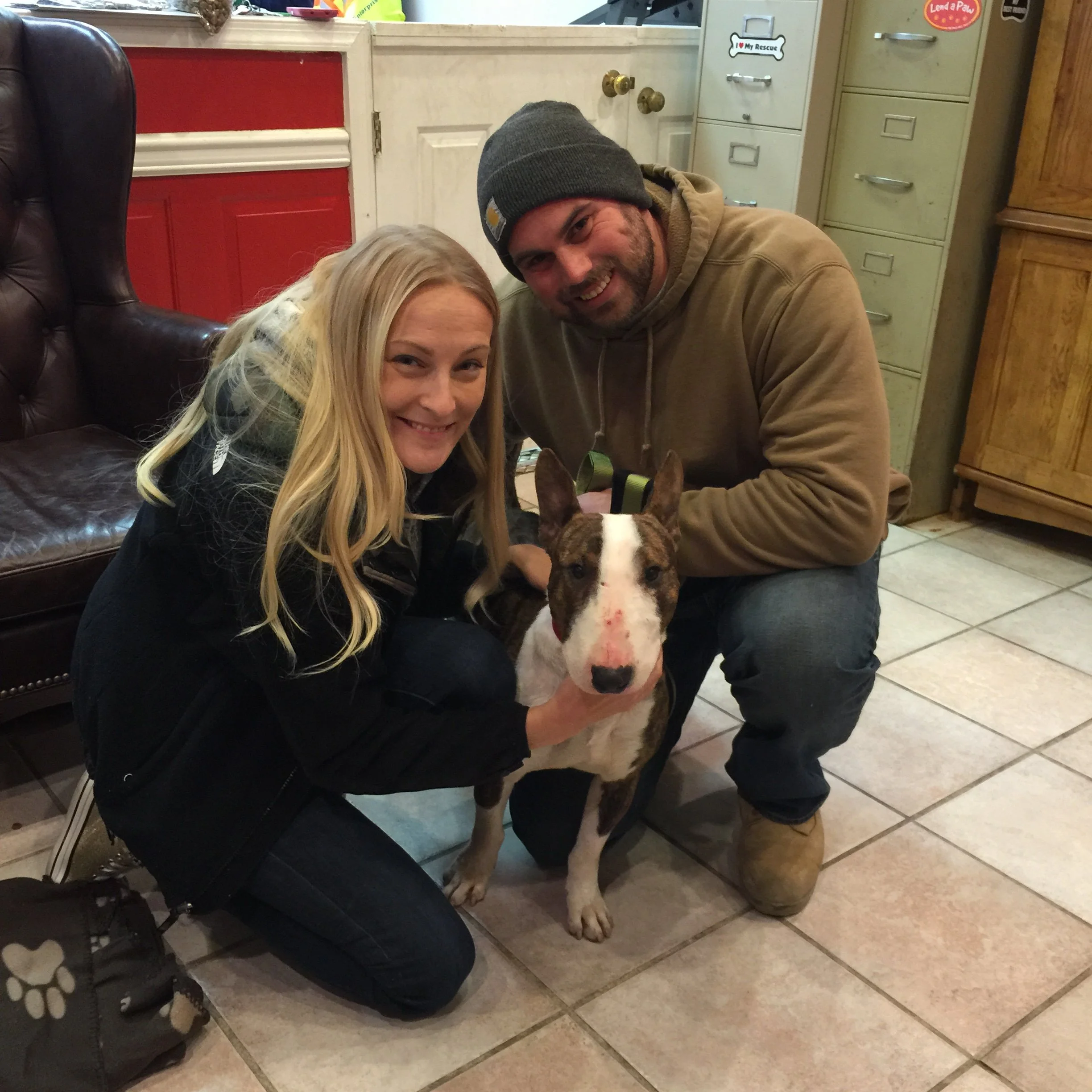 A woman and a man are smiling while sitting on the floor, hugging a dog. The man is wearing a gray beanie and a tan hoodie. The woman has long blonde hair. The background shows a red painted door, a beige cabinet with some stickers, and tiled floorin