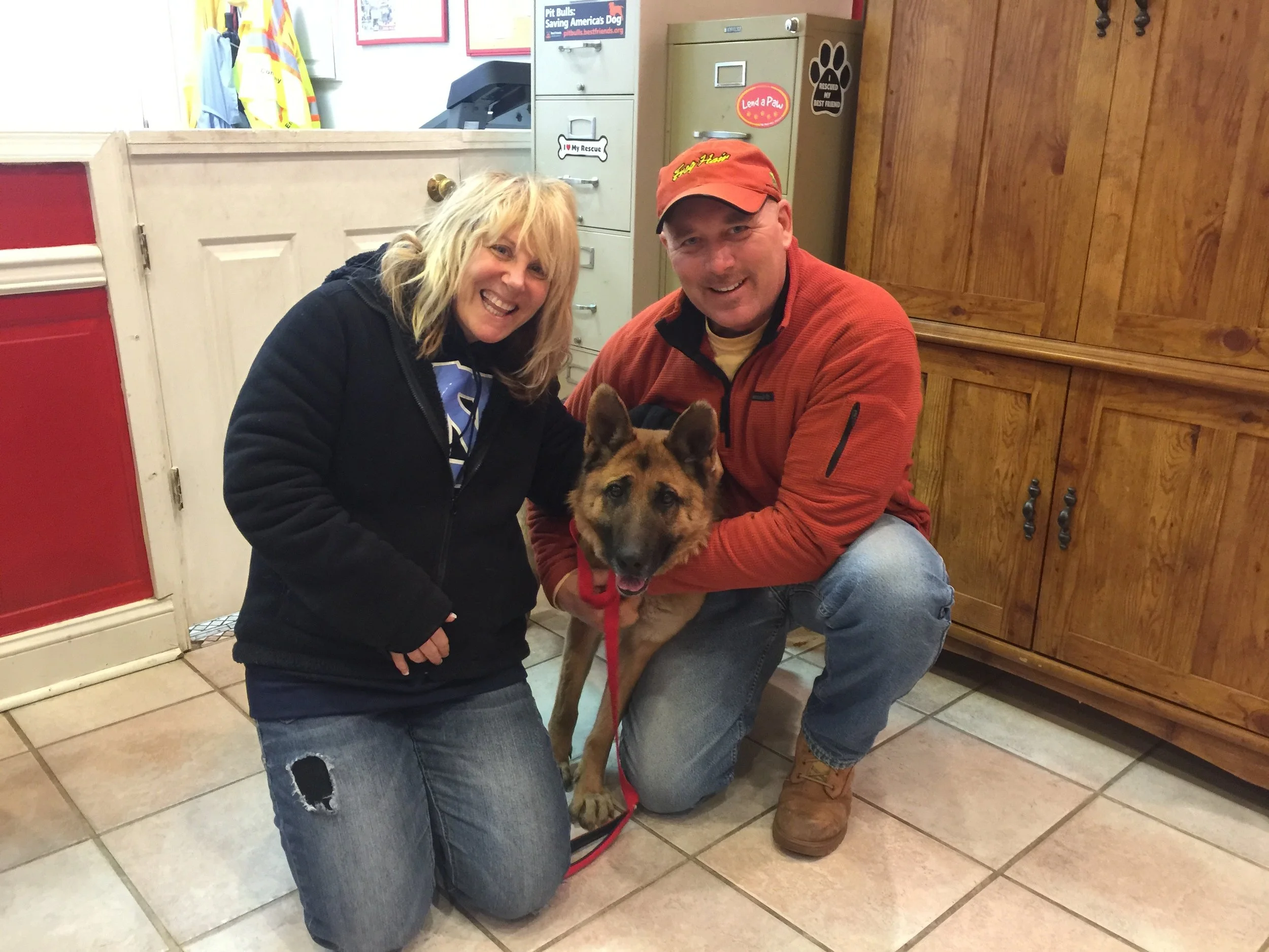 A woman and a man crouching next to a German Shepherd dog inside a room. The woman is wearing a black jacket, blue jeans with a ripped knee, and has blonde hair. The man is wearing a red cap, an orange jacket, and blue jeans. The room features wood p