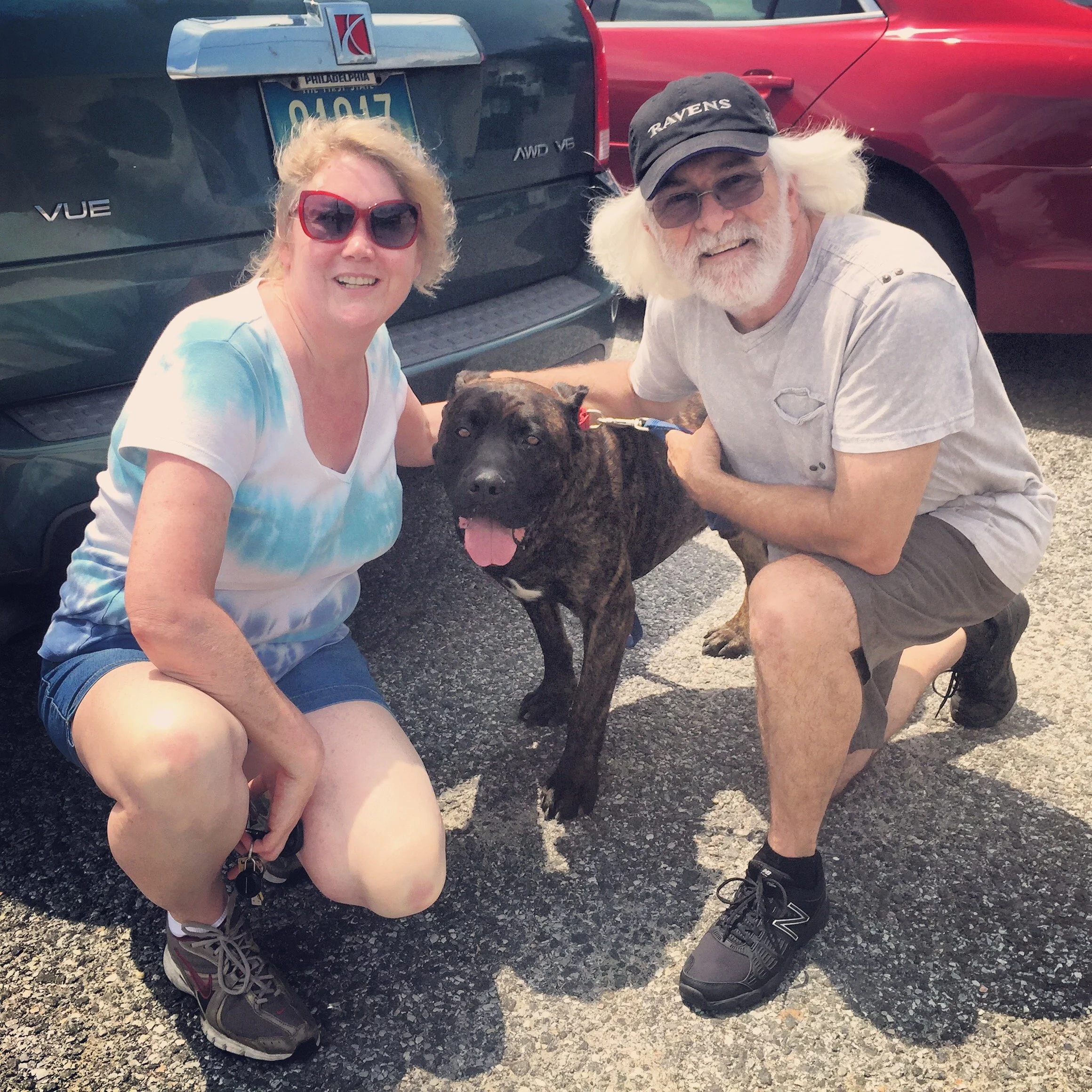 Two smiling people, a woman with blonde hair wearing sunglasses and a white shirt and a man with white hair, beard, wearing a baseball cap that says Ravens, and sunglasses, are squatting and holding a black dog with a red collar between them on a par