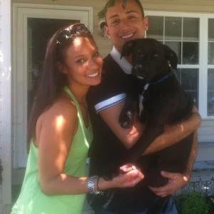 A smiling couple holding a black dog outdoors near a house with large windows.