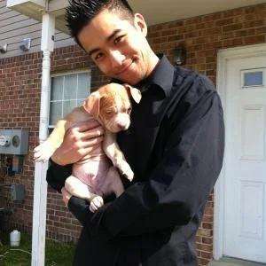 A young man smiling and holding a small dog in his arms outside a brick house.