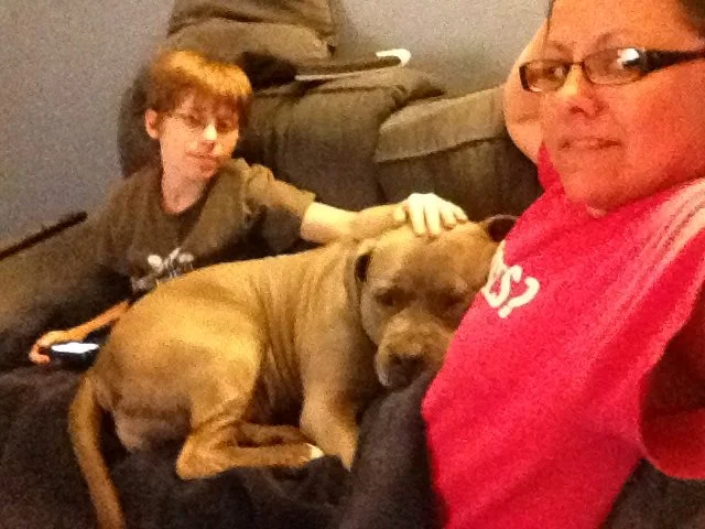 A woman and a young boy sitting on a couch with a large brown dog. The woman is smiling and taking a selfie, while the boy is touching the dog and looking at it. The dog is resting with its head on the woman's lap.