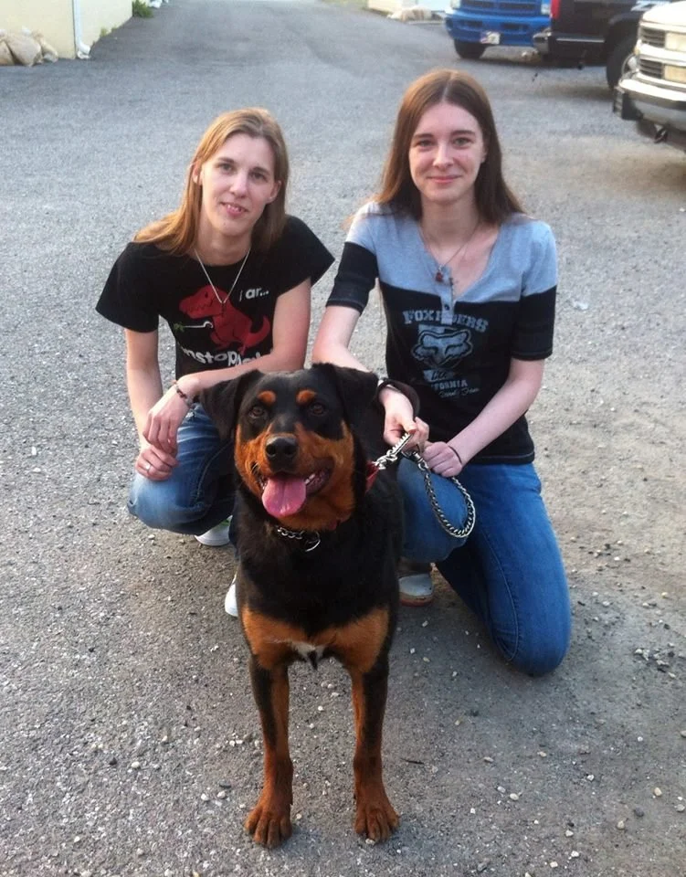 Two young women kneeling on a gravel driveway with a large Rottweiler mix dog, all smiling and looking at the camera.