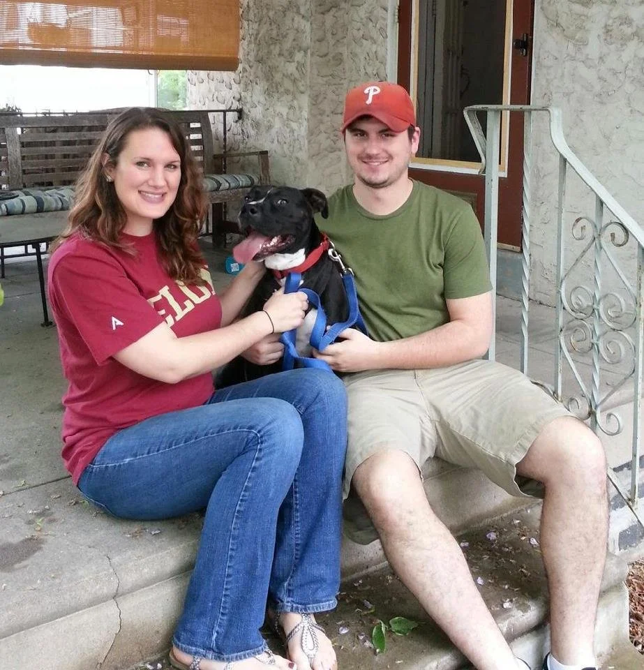 A woman and a man sitting on a porch steps with a black dog between them. The woman is holding the dog's chest and smiling, wearing a red t-shirt and jeans. The man, wearing a green t-shirt, beige shorts, and a red baseball cap, is holding the dog’s 