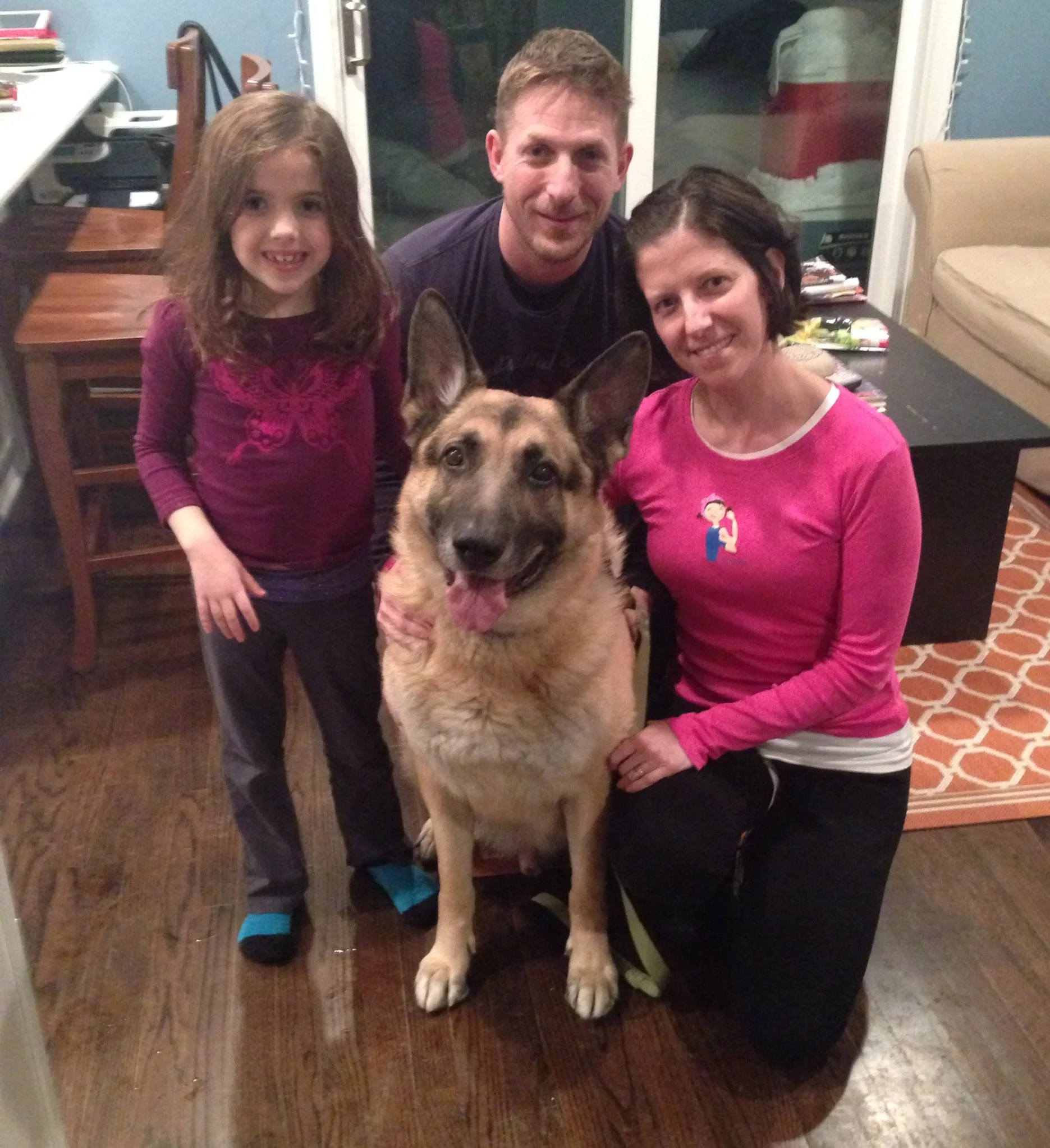 A group of three people and a German Shepherd dog inside a room, with two women, one man, and a girl. The girl is smiling, the man is looking at the camera, and the woman is sitting with the dog. The room has a table with magazines and books, a beige