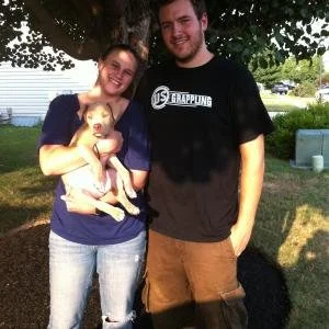 A woman and a man standing outdoors under a tree, smiling. The woman is holding a small dog, and the man is wearing a black T-shirt and khaki shorts.