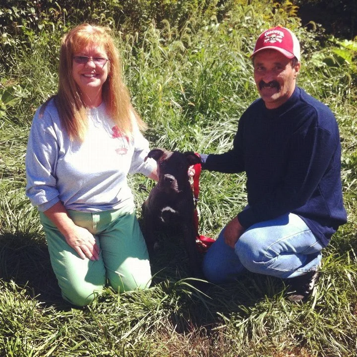 A woman and a man are kneeling outdoors in tall grass, smiling at the camera, with a black goat between them. The woman has long red hair and is wearing glasses, a white shirt, and light green pants. The man has dark hair, a mustache, and is wearing 