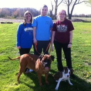 Three people standing outdoors on grass with two dogs, a large one and a smaller one, on leashes.