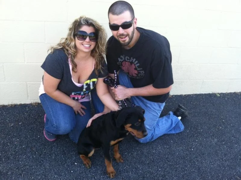 A smiling woman and a man in sunglasses pose with a puppy against a beige wall, kneeling on the pavement.