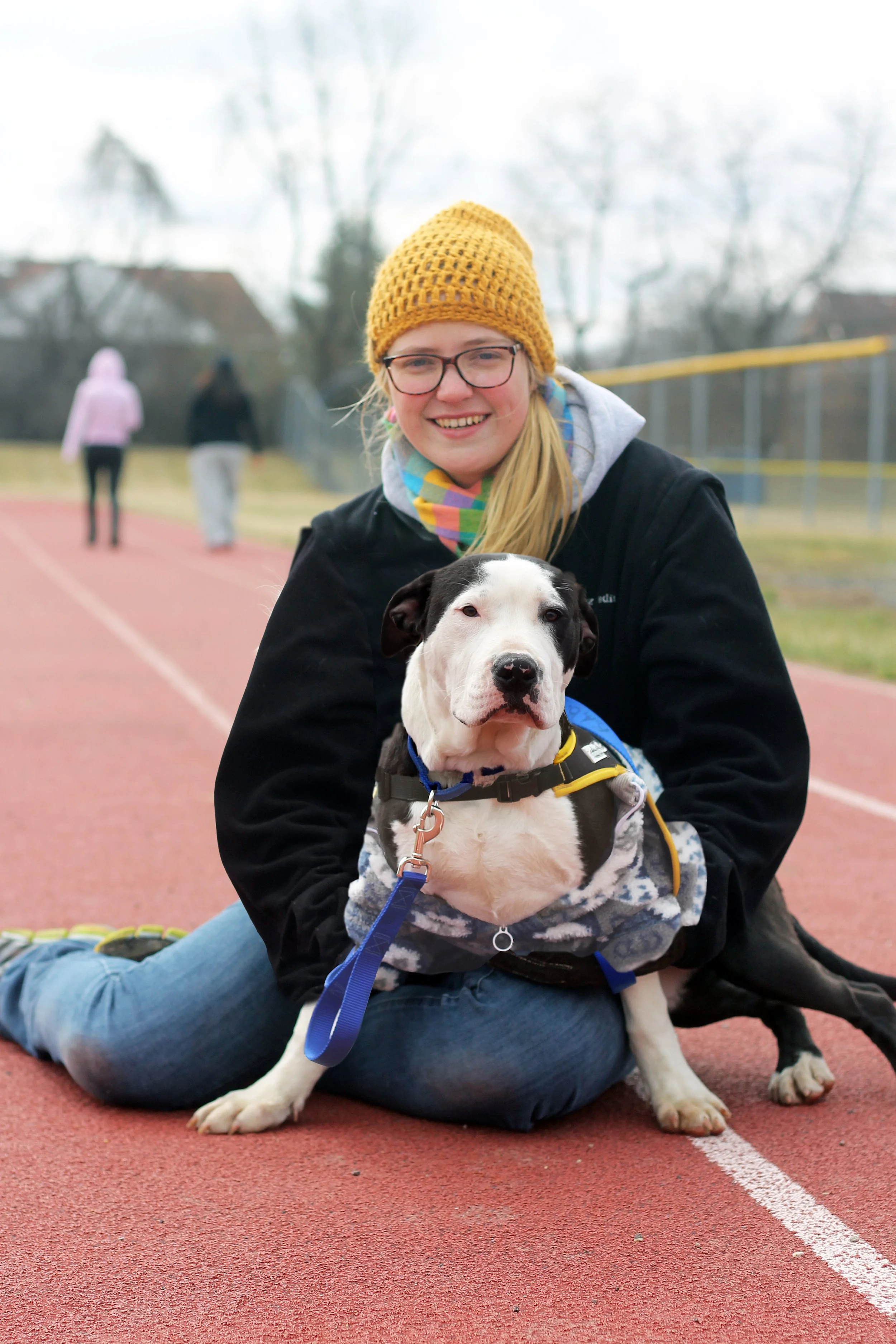 A smiling young woman with glasses, a yellow knit hat, and a gray hoodie, sitting on a running track with her black and white puppy, which has a blue leash, in her lap. The background shows a few people walking on the track and leafless trees.