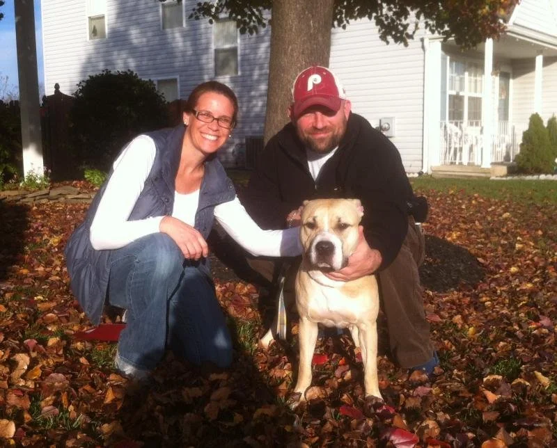 A woman and a man kneeling on autumn leaves outdoors with a large dog between them, smiling at the camera. The woman is wearing glasses, a white long-sleeve shirt, and a dark vest. The man is wearing a red baseball cap, black hoodie, and brown pants.
