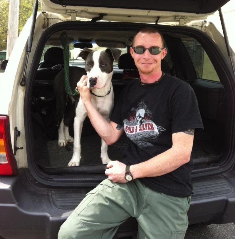 A man sitting on the back of an open SUV trunk, smiling and holding a dog with a black and white coat, inside a suburban neighborhood during the daytime.