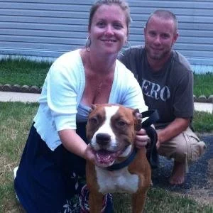 A woman and a man outdoors with a brown and white dog, all smiling.