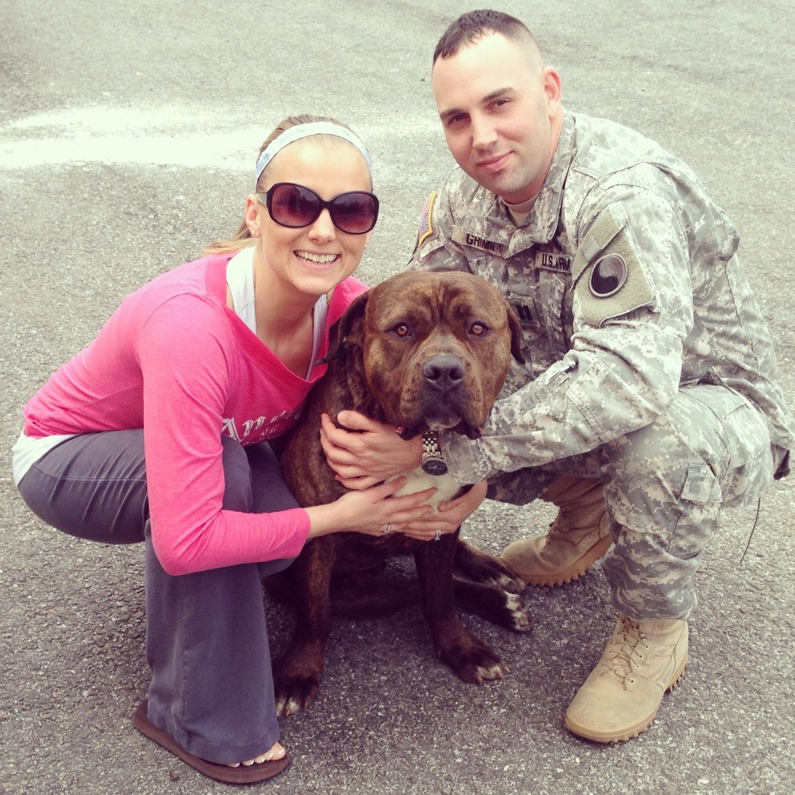 A woman and a man are crouching on a gray pavement, smiling and holding a large brown dog between them. The woman is wearing a pink long sleeve shirt, sunglasses, and a white headband. The man is in U.S. Army camouflage uniform with patches and a nam