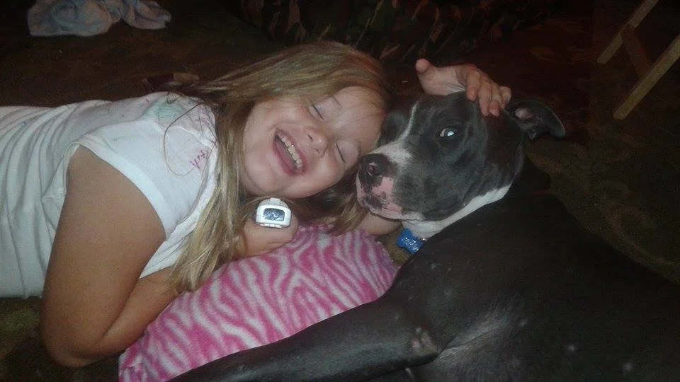 Girl lying on her side with a big smile, hugging a black and white dog with a serious expression, on a pink and white striped blanket.