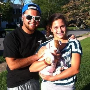 A smiling man and woman outdoors holding a puppy together on a sunny day.