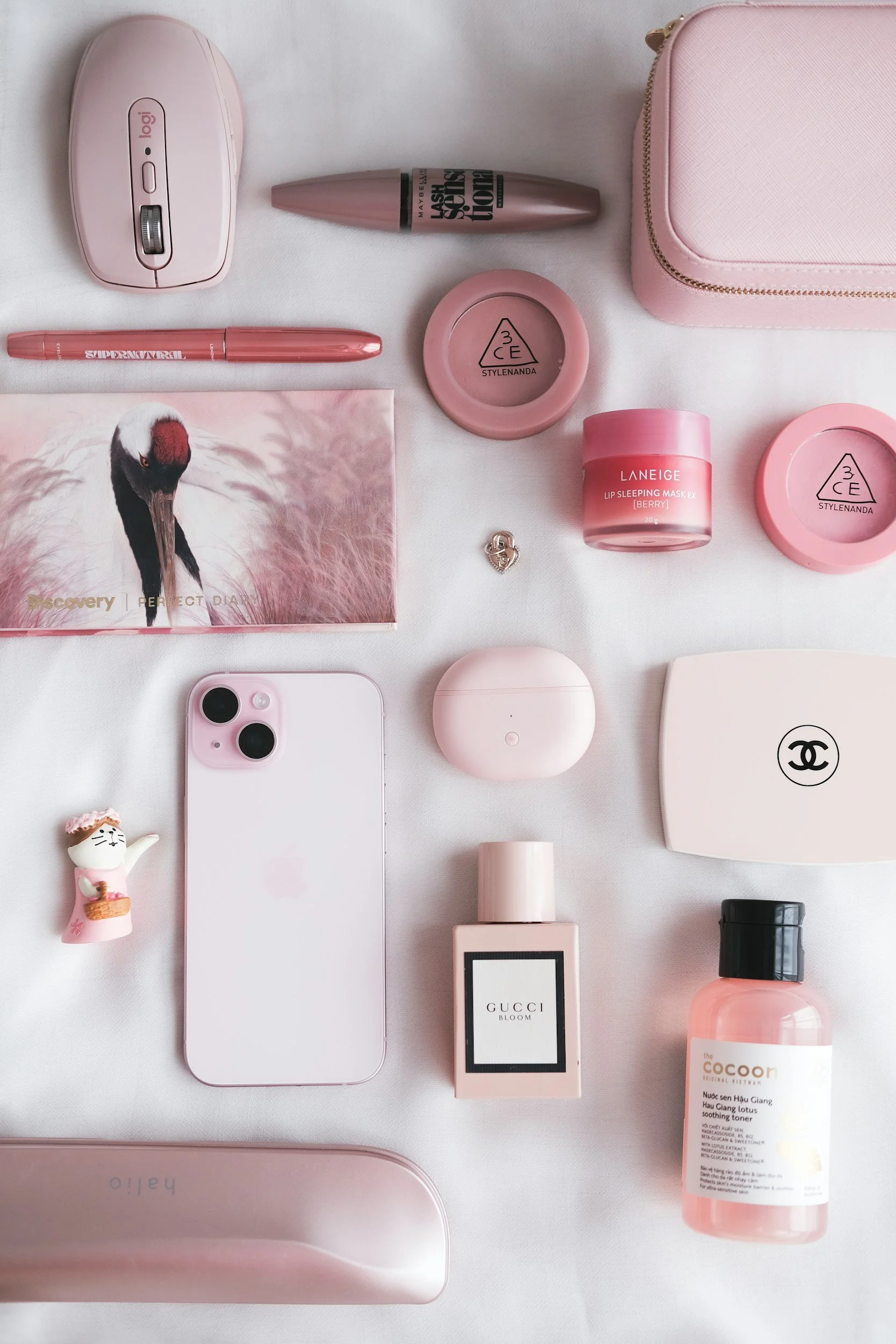 Flat lay of pink and white beauty and tech accessories, including an iPhone, Gucci perfume, Laneige lip sleeping mask, Chanel compact mirror, and various skincare bottles and containers, arranged on a white surface.