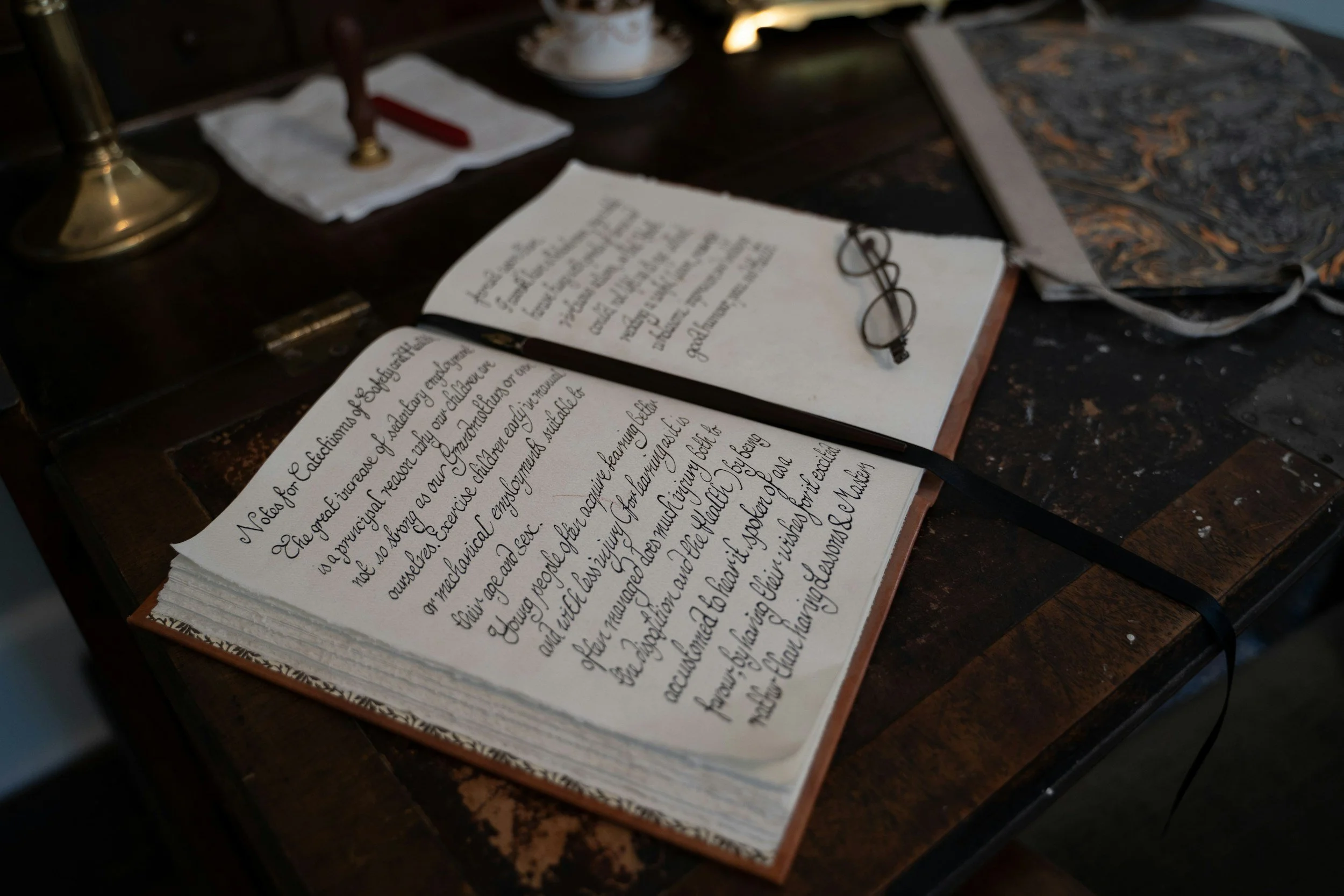 An open notebook on a wooden desk with handwritten notes in cursive, a pair of reading glasses resting on the pages, a brass desk lamp, and a patterned bag in the background.