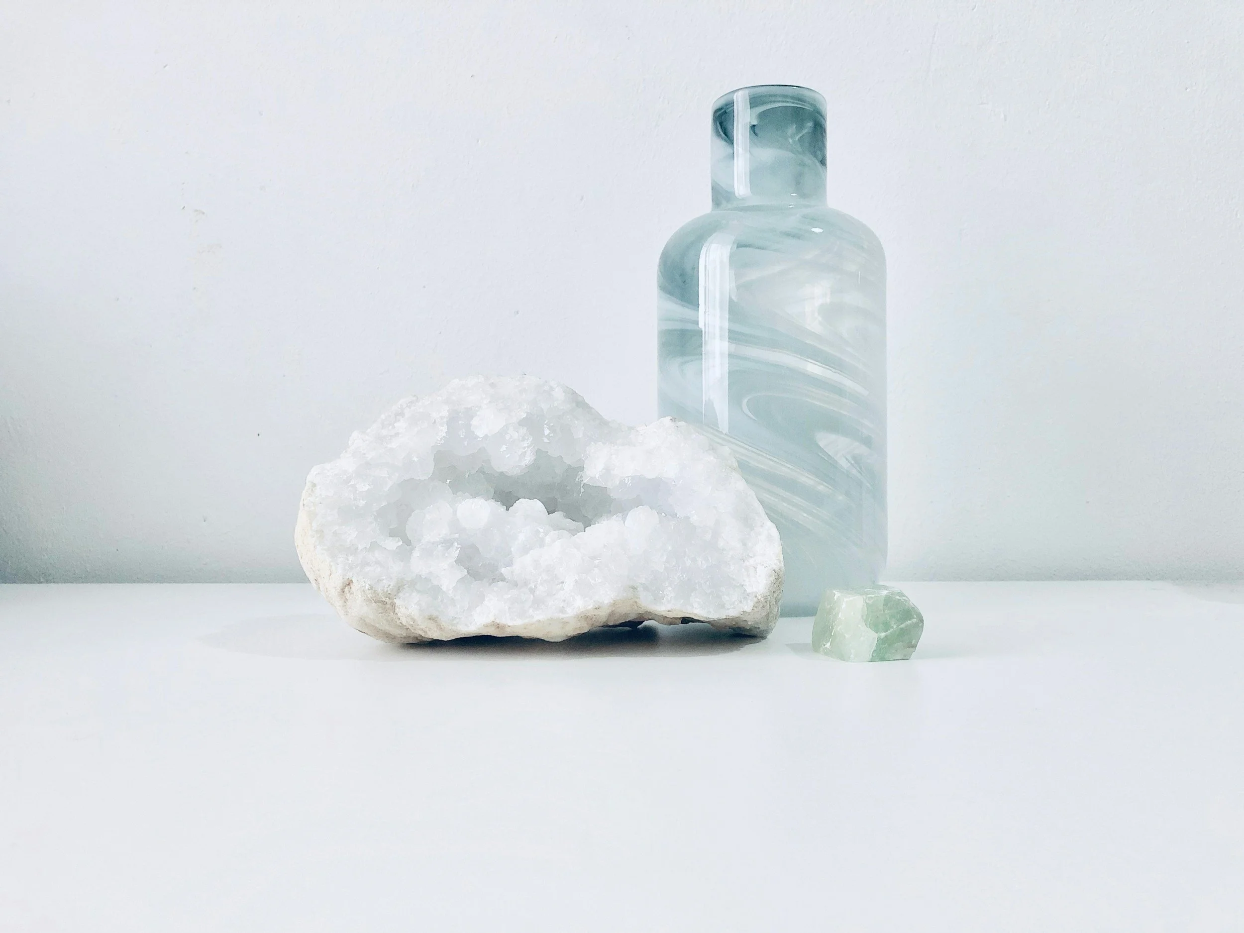 A tall, cylindrical glass bottle with a swirling blue and white marble pattern, placed on a white surface with a lack of background details. Next to it, there is a rough, white, textured stone and a small, greenish, semi-translucent cube, likely a piece of mineral or crystal.