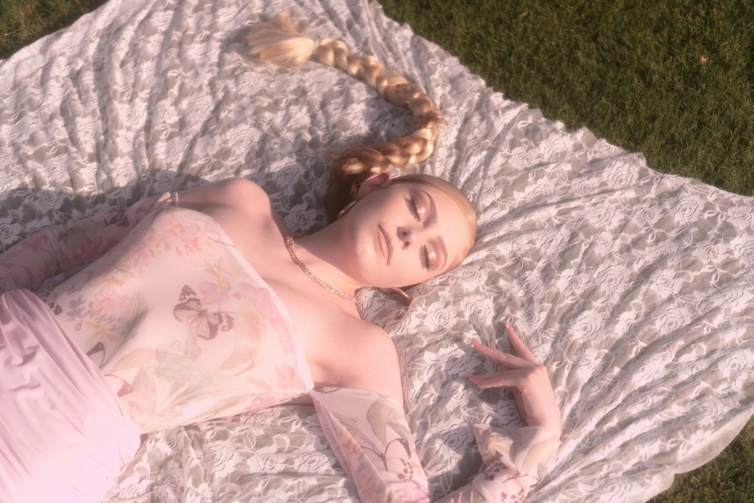 A woman with long braid and makeup, wearing a pink floral dress, lying on lace blanket outdoors with grass visible.