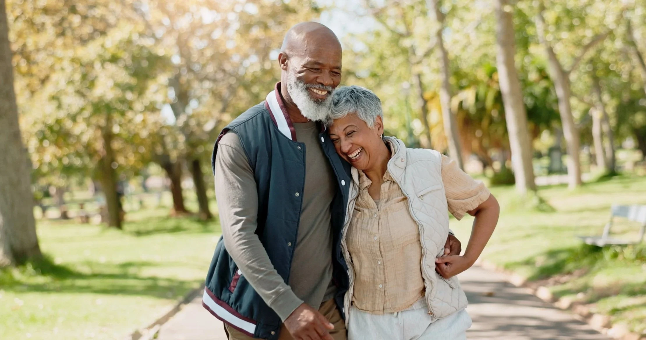 An elderly couple walking together in a park with trees and green grass, smiling and happy.